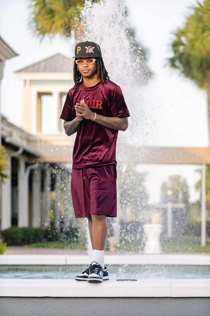 Man wearing burgundy velour short set from summer collection standing near fountain outdoors